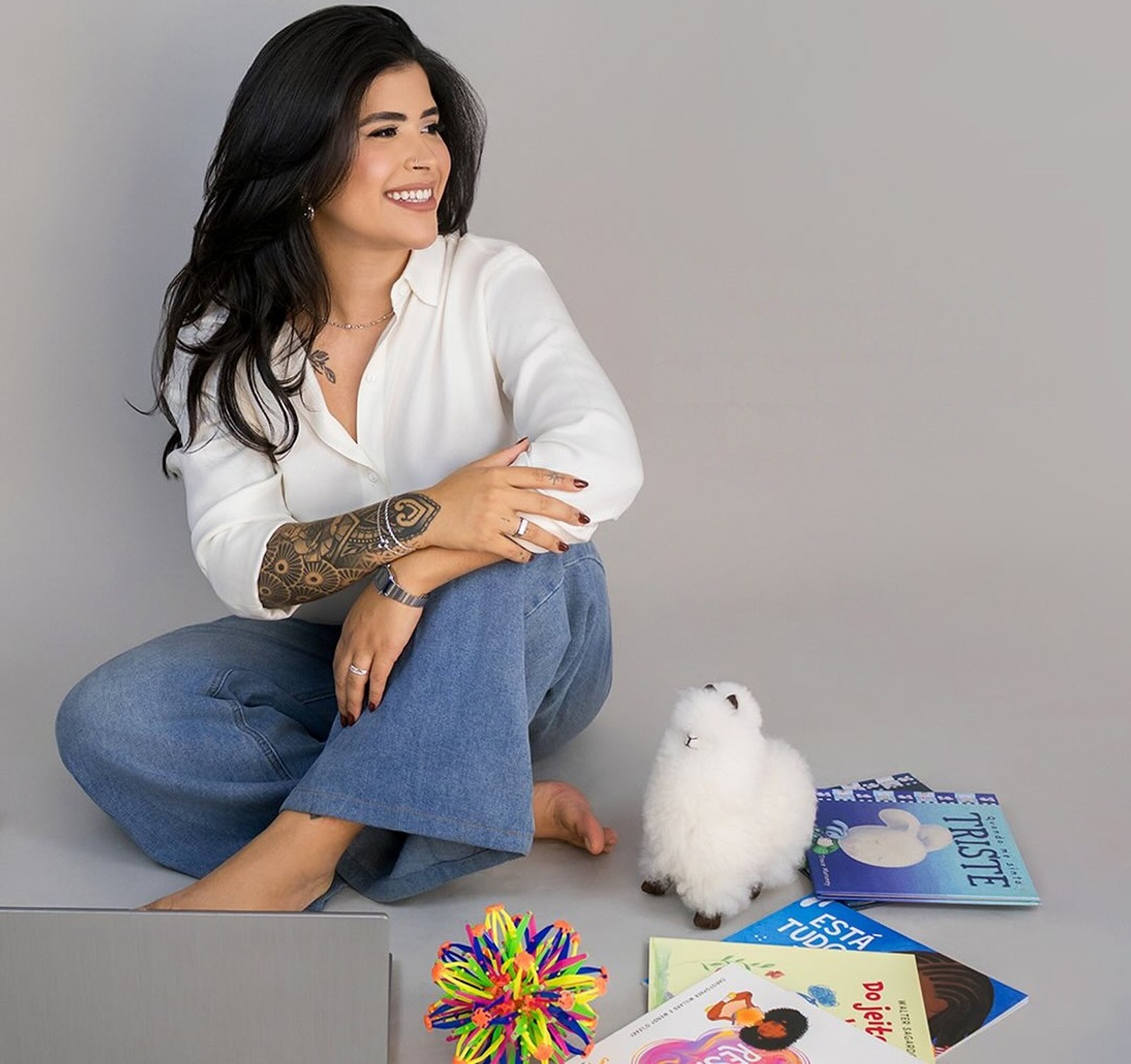 Fernanda sentada no chão, sorrindo, com livros infantis e brinquedos ao lado